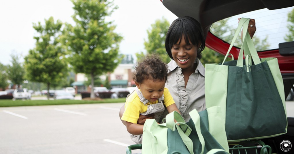 A toddler curiously peeks into a grocery bag as his mother puts them in the trunk of their car