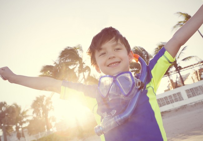 A young boy, wearing a scuba diving outfit, throws his arms up happily at the beach