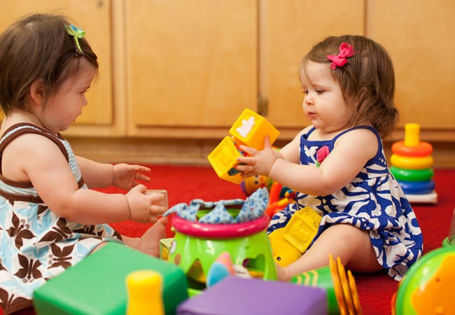 Two little toddler girls, surrounded by different toys, play with blocks at a day care center