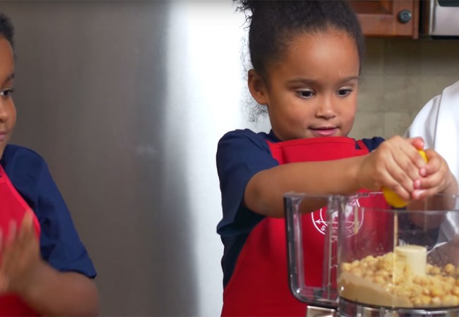Little girl squeezing lemon juice into a food processor full of chickpeas to make hummus