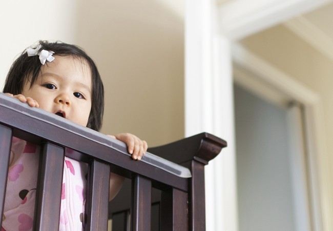 An adorable little girl stands inside her baby crib and peers out over the railing.