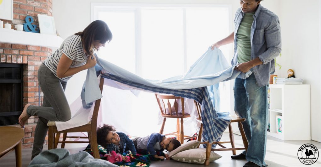 Parents build a blanket fort for their children as they smile happily sitting under it