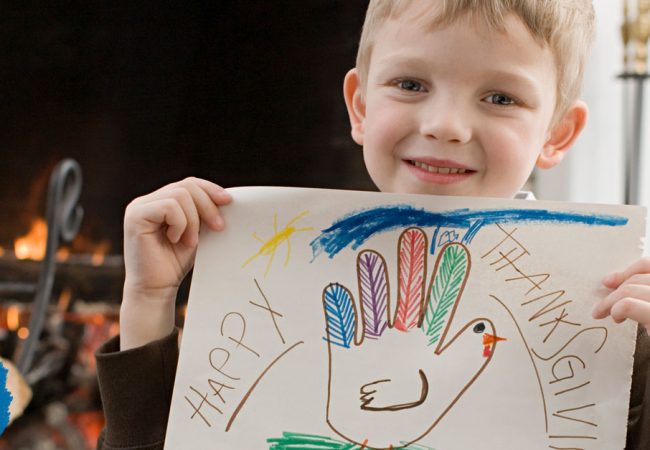 A young boy holds up a drawing of a hand-shaped turkey with happy thanksgiving written around it