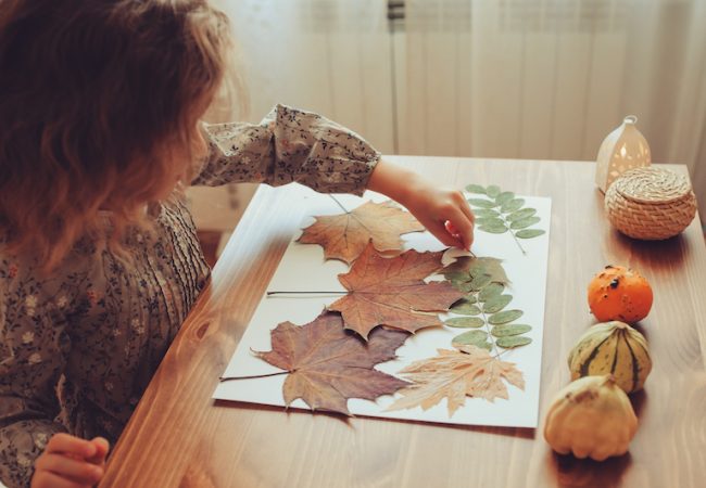 Little girl sitting at table creates fall craft with leaves and construction paper