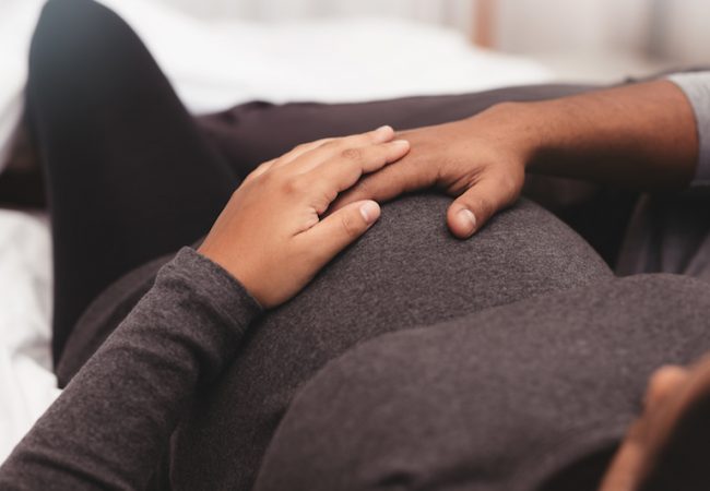 Close up of pregnant couple lying on bed at home. Focus on hands of man and woman on a pregnant belly.
