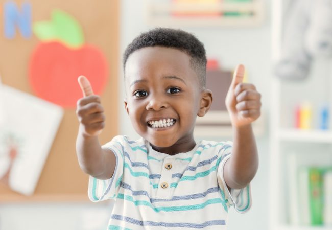Little boy at preschool smiles and gives thumbs up to camera on back to school