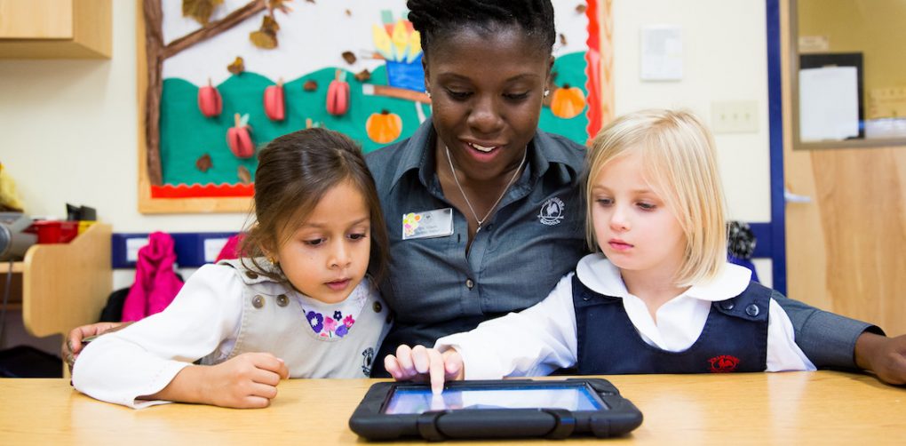Primrose teacher sits at table and helps two girl preschoolers use tablet