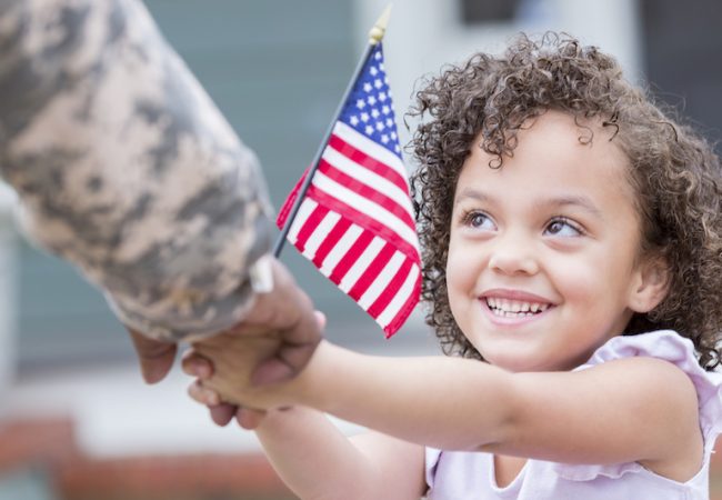 Little girl holds American flag and hand of woman in military uniform for Veterans Day