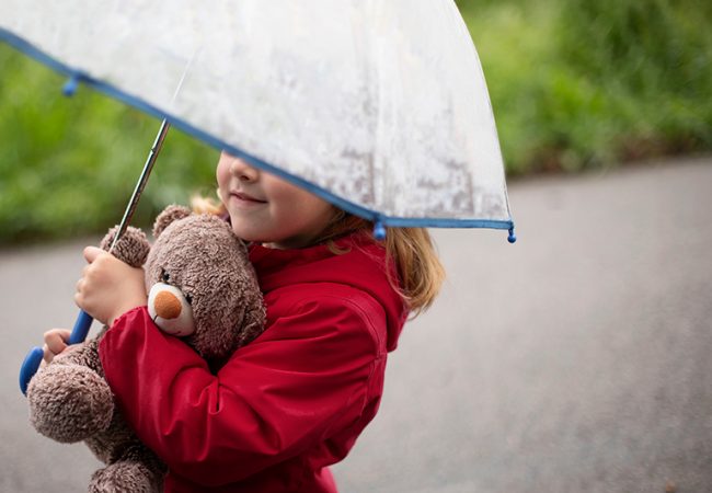 Girl cuddling teddy bear and walking under and umbrella in the rain.